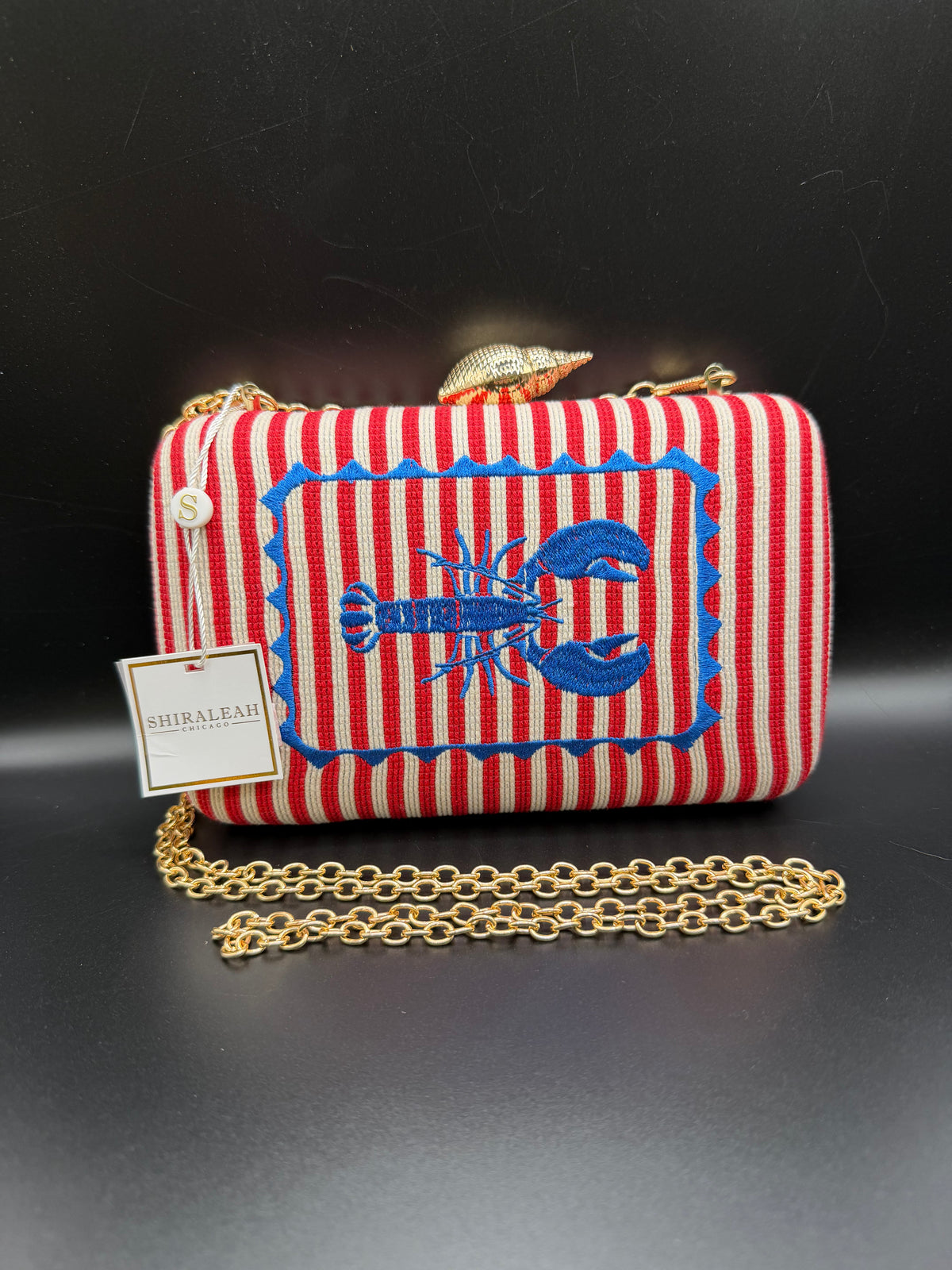 The selected item is the Shiraleah Marina Red Striped Lobster Minaudière Clutch.This nautical-inspired evening bag features a structured box silhouette dressed in bold red and white vertical stripes. It is accented by a prominent, bright blue embroidered